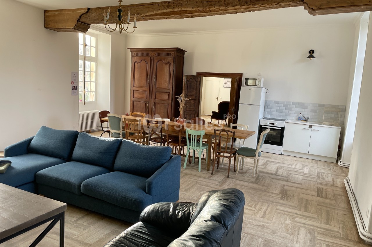 Salle de séjour lumineuse avec canapé, table à manger, armoire en bois, cuisine équipée et poutre apparente.