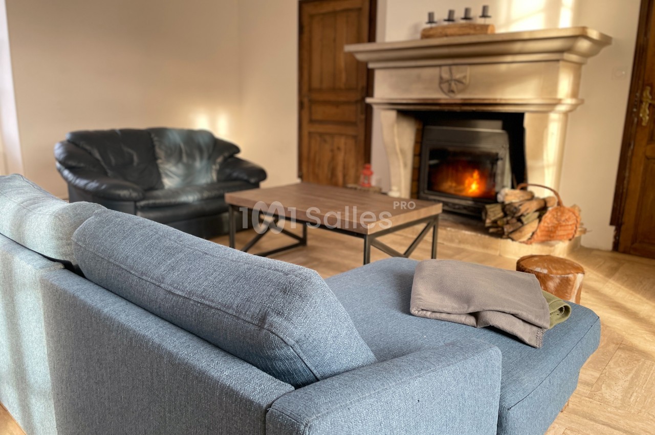 Salon chaleureux avec canapé bleu, fauteuil en cuir noir, table basse et cheminée allumée dans un décor lumineux.