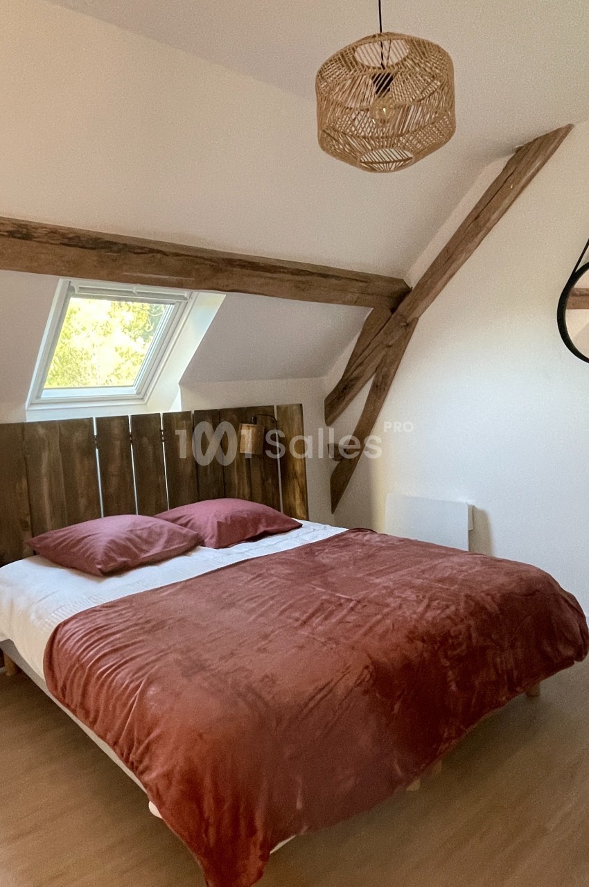 Chambre lumineuse avec lit double, tête de lit en bois, poutres apparentes et fenêtre de toit.