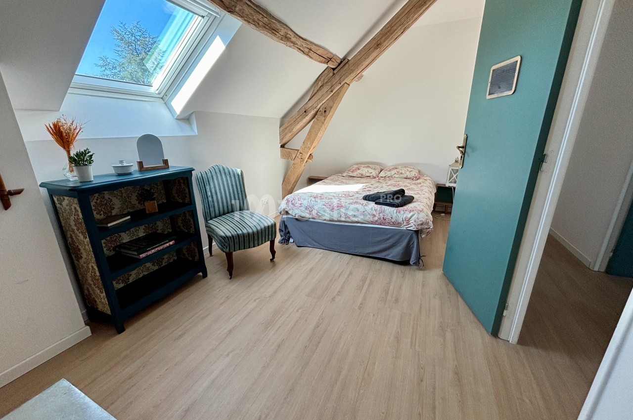 Chambre lumineuse avec lit double, poutres apparentes, fauteuil, étagère et fenêtre de toit.