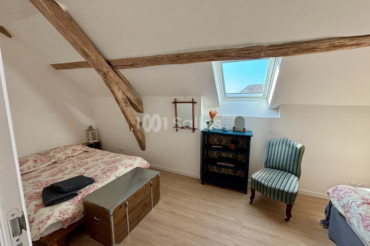 Chambre mansardée avec deux lits simples, poutres apparentes, fenêtre de toit et mobilier simple.