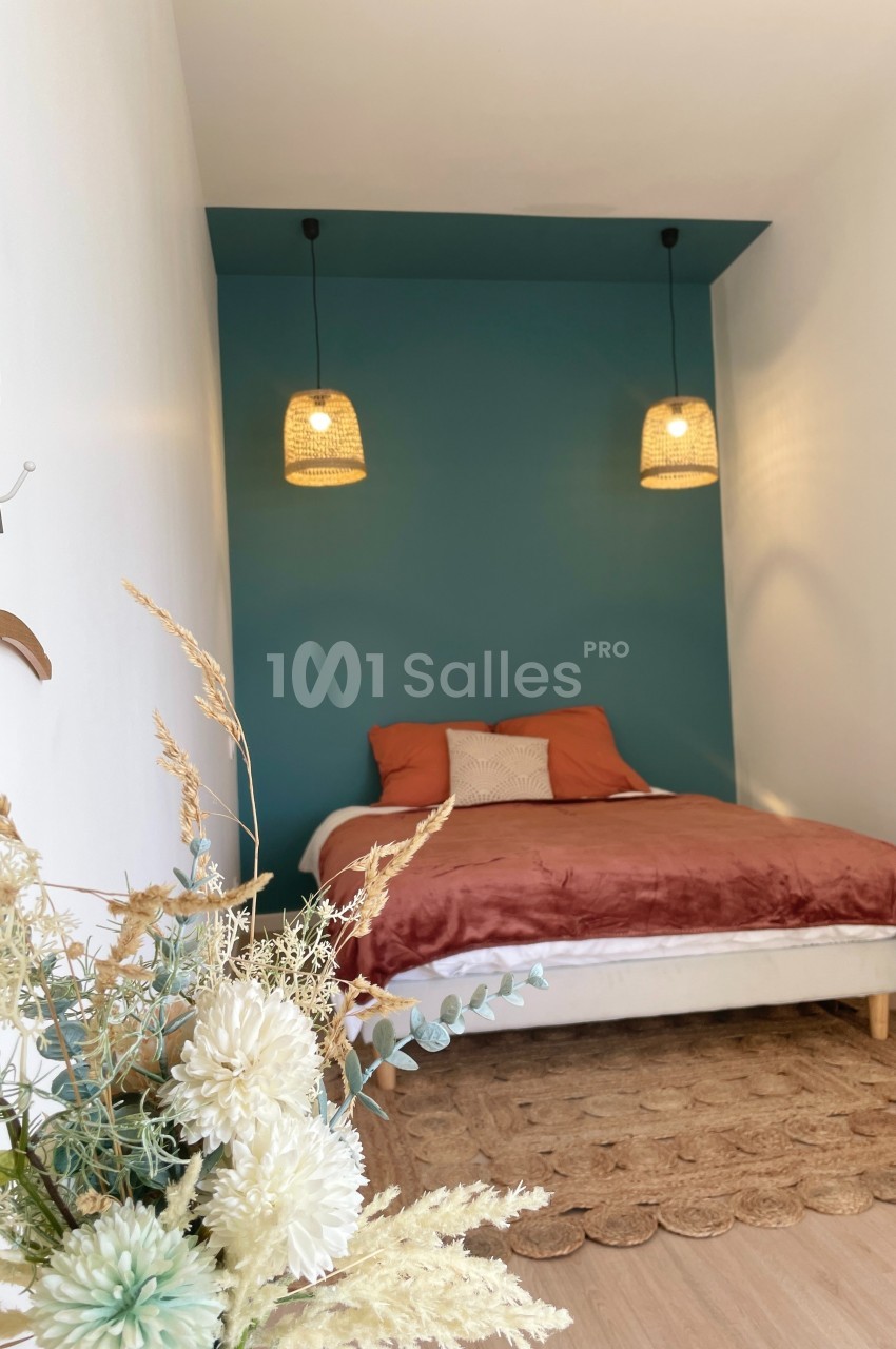 Lit double avec coussins et couverture dans une chambre minimaliste, mur bleu, suspensions en rotin et décoration florale.