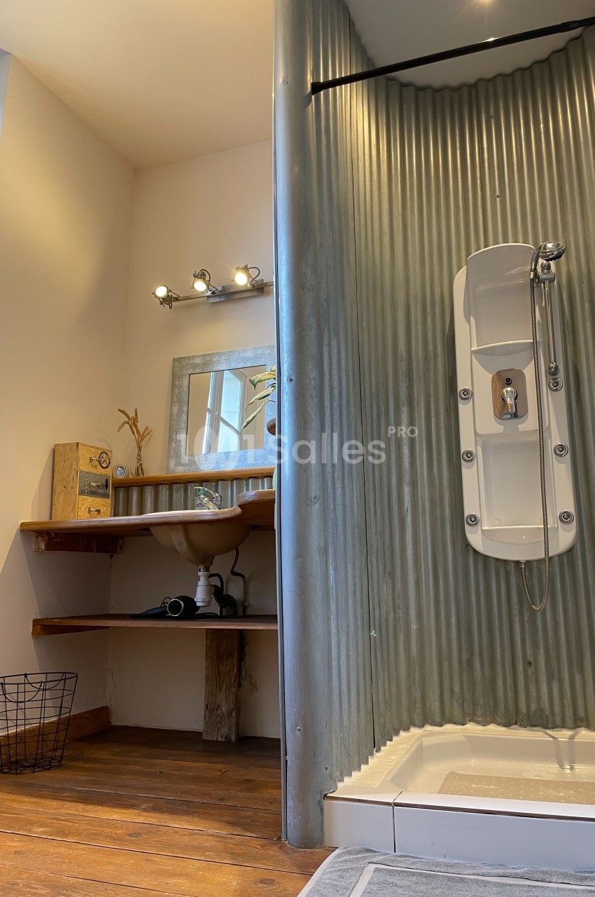 Salle de bain avec douche en tôle ondulée, lavabo sur planche en bois et miroir encadré, éclairage au-dessus.