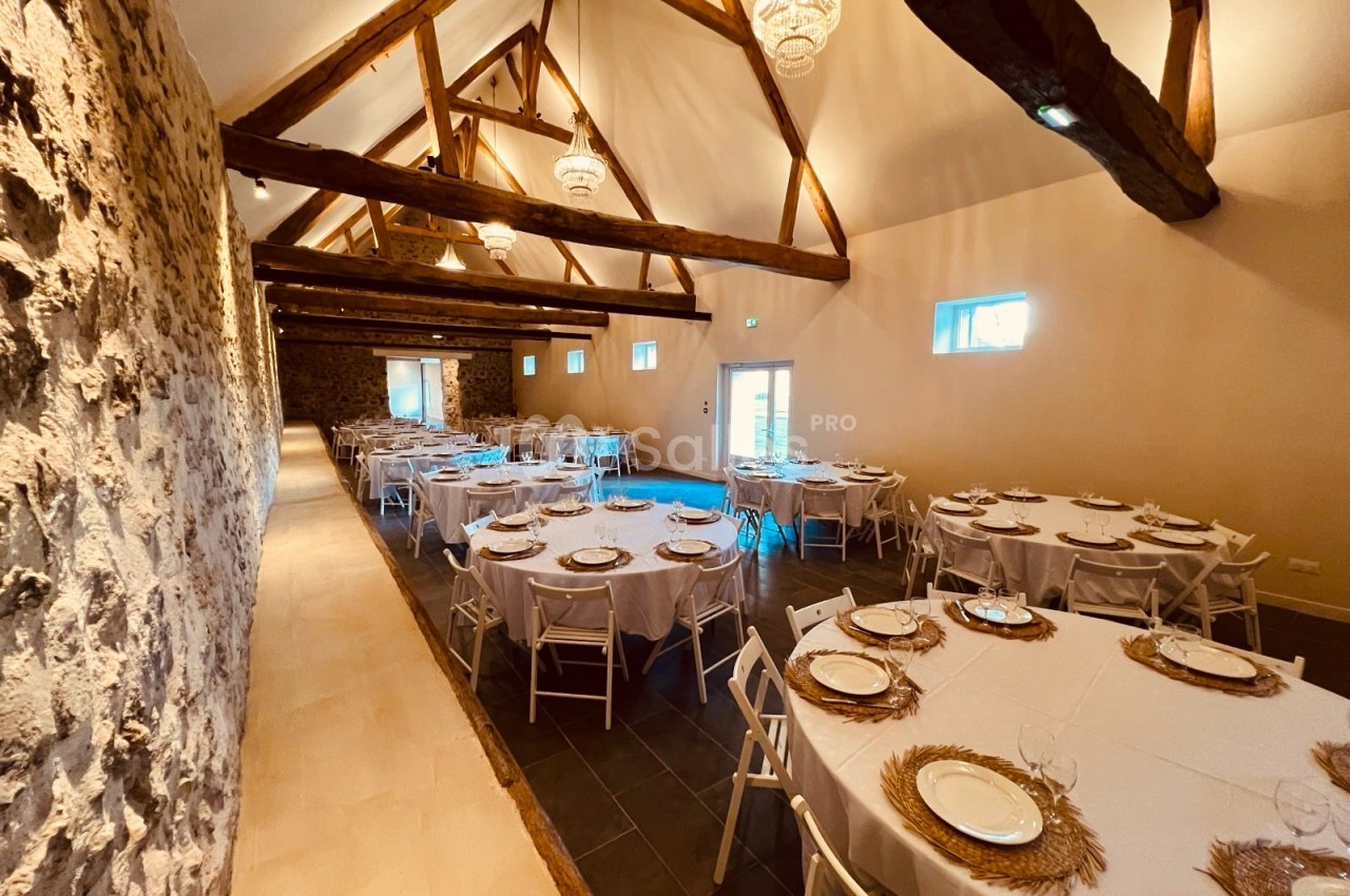 Salle de réception avec tables rondes dressées, murs en pierre apparente et poutres en bois au plafond.