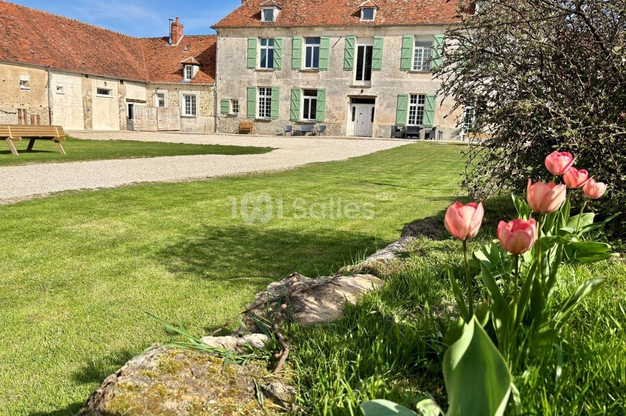 Jardin verdoyant avec des tulipes roses au premier plan et une maison ancienne en pierre avec volets verts en arrière-plan.