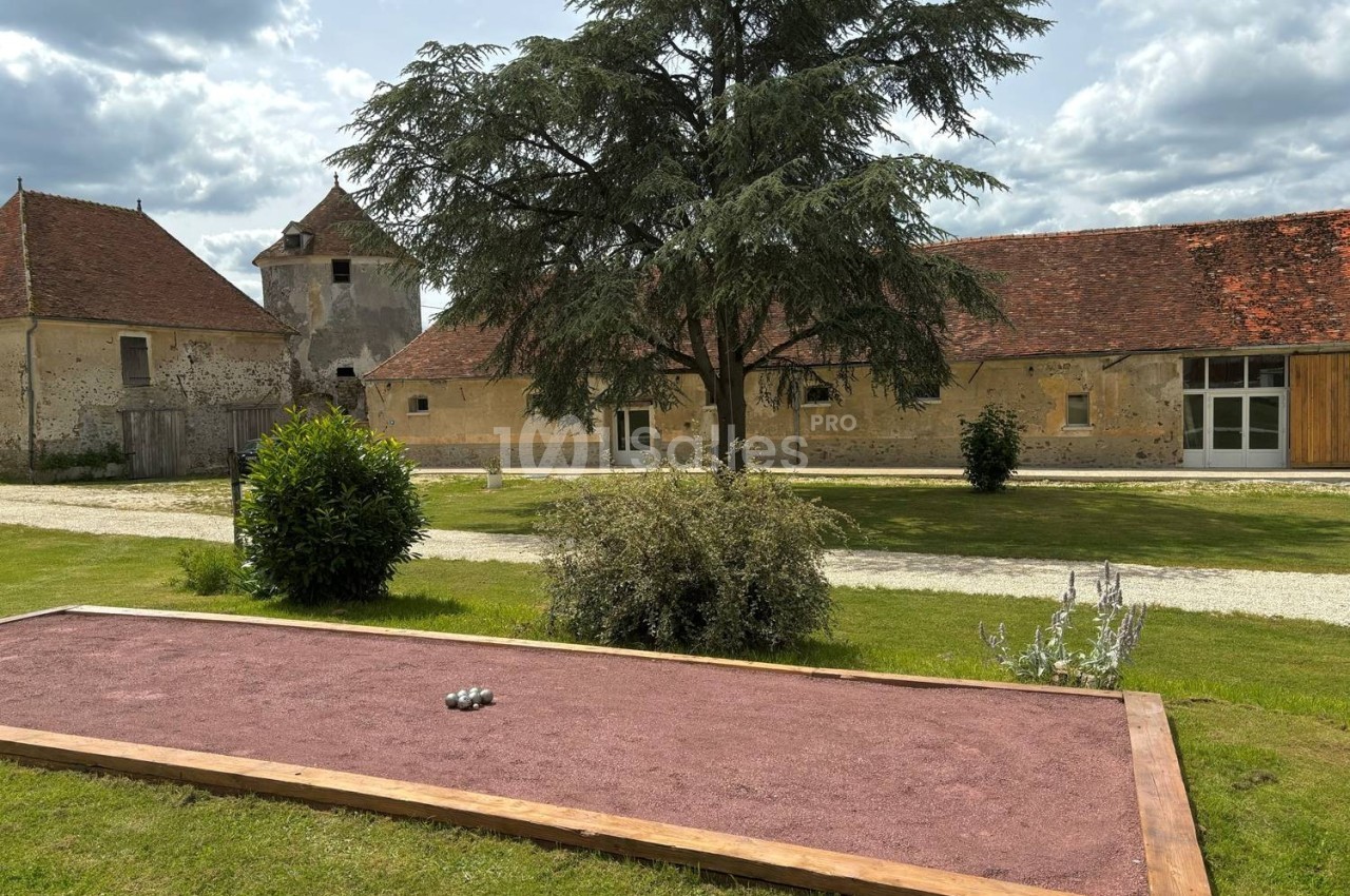 Terrain de pétanque en gravier rouge devant un grand arbre et des bâtiments anciens en pierre sous un ciel nuageux.