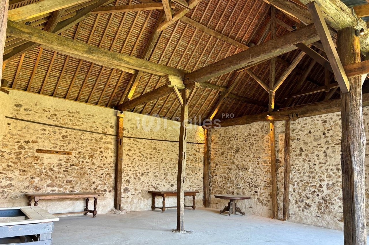 Intérieur d'une grange en pierre avec charpente en bois apparente et quelques tables en bois contre les murs.