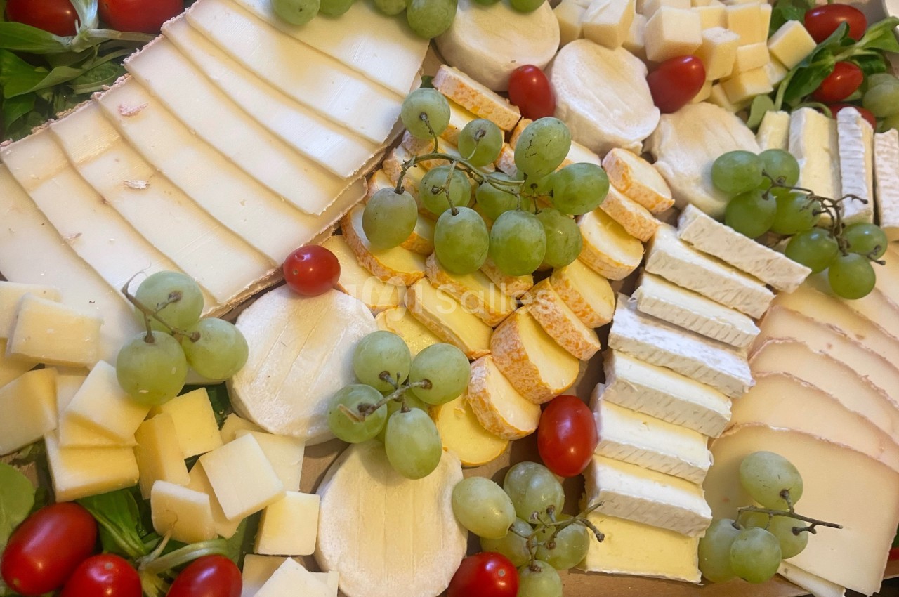 Plateau de fromages variés avec raisins verts, tomates cerises et feuilles de salade en décoration.