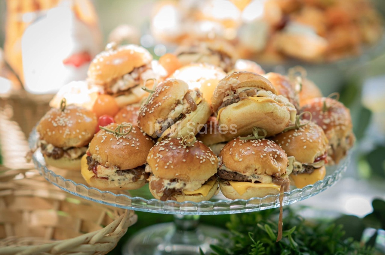 Plateau de mini-burgers garnis, disposés sur un présentoir en verre, entouré d'éléments décoratifs.
