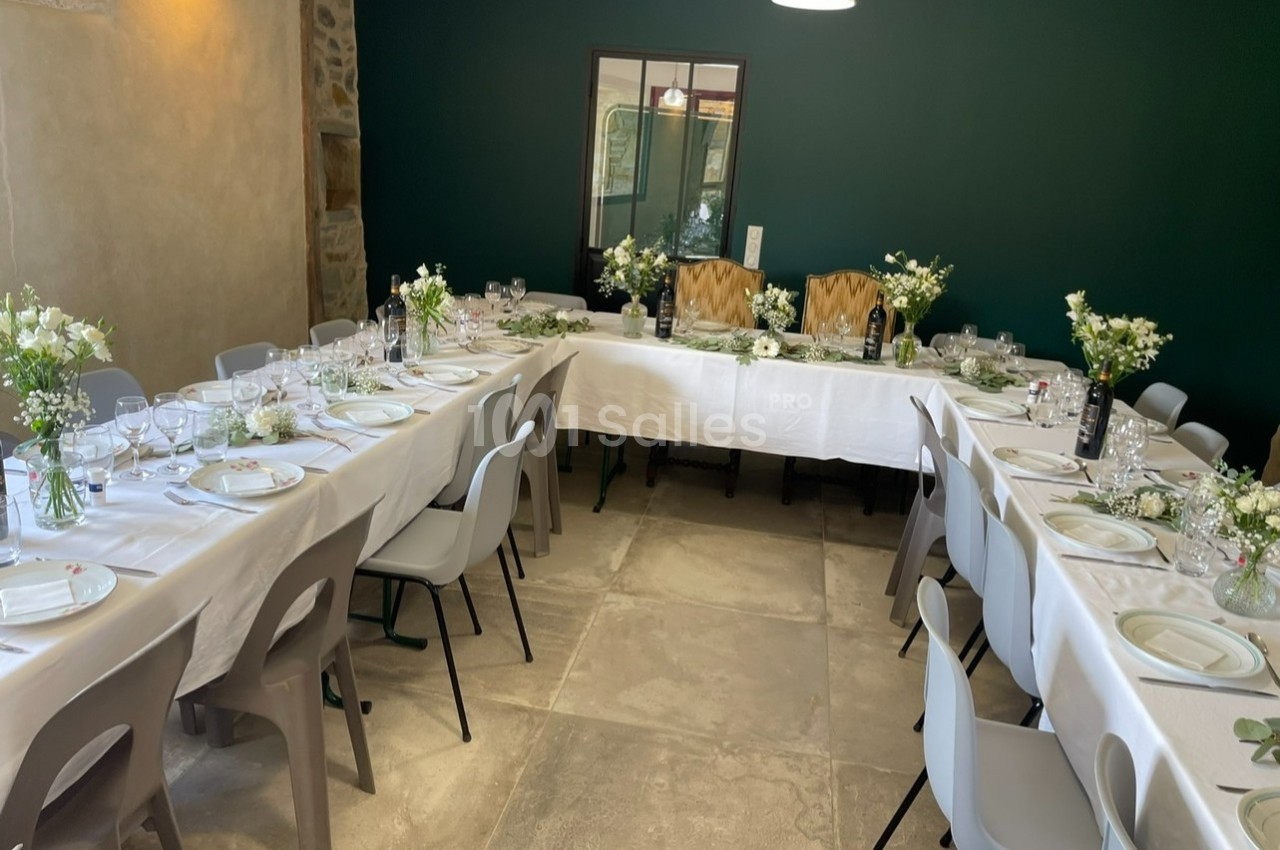 Salle décorée pour un repas, avec tables en U dressées de nappes blanches, vaisselle et bouquets de fleurs.