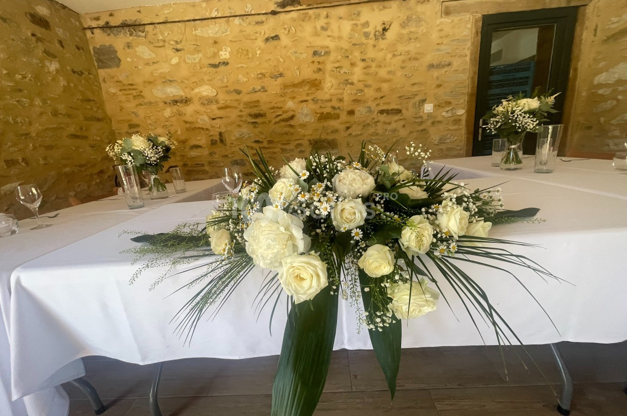 Centre de table floral avec roses blanches et feuillage, disposé sur une nappe blanche dans une salle en pierre.