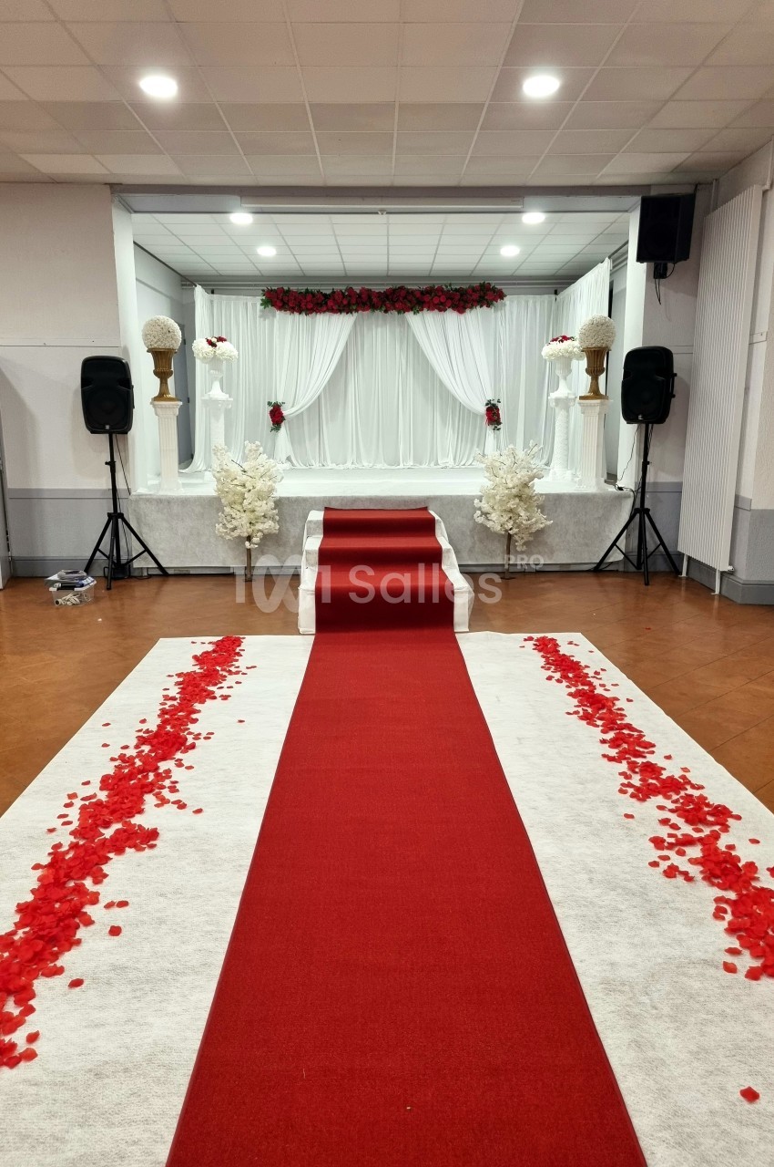 Allée rouge bordée de pétales de roses menant à une scène décorée de fleurs blanches et rouges dans une salle de réception.