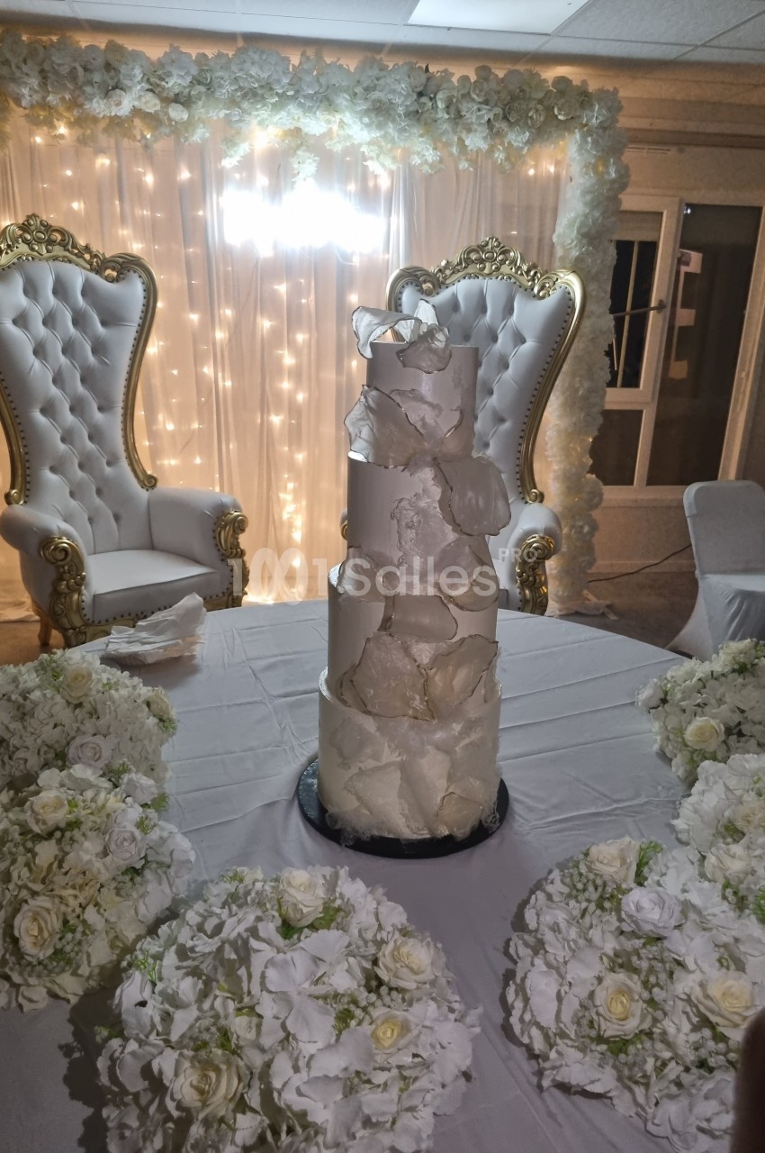 Gâteau de mariage à plusieurs étages décoré de blanc, entouré de bouquets de fleurs, avec deux fauteuils ornés en arrière…