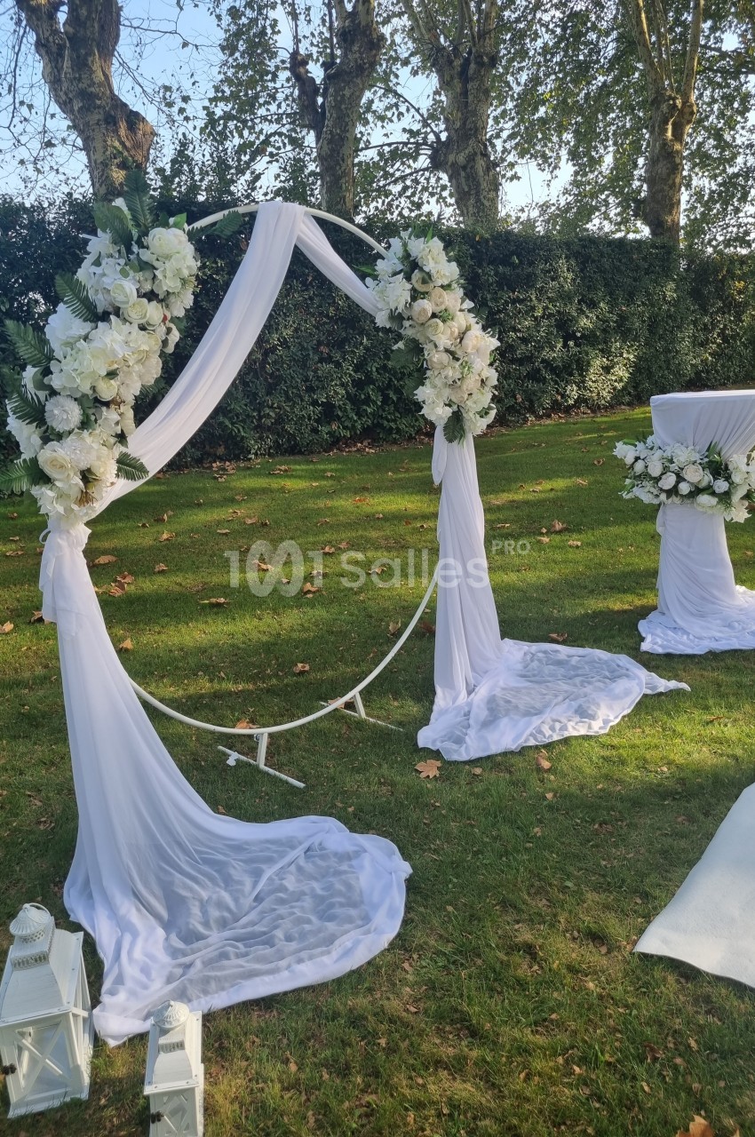 Arche de mariage circulaire décorée de fleurs blanches et de voilages, installée sur une pelouse avec lanternes blanches.