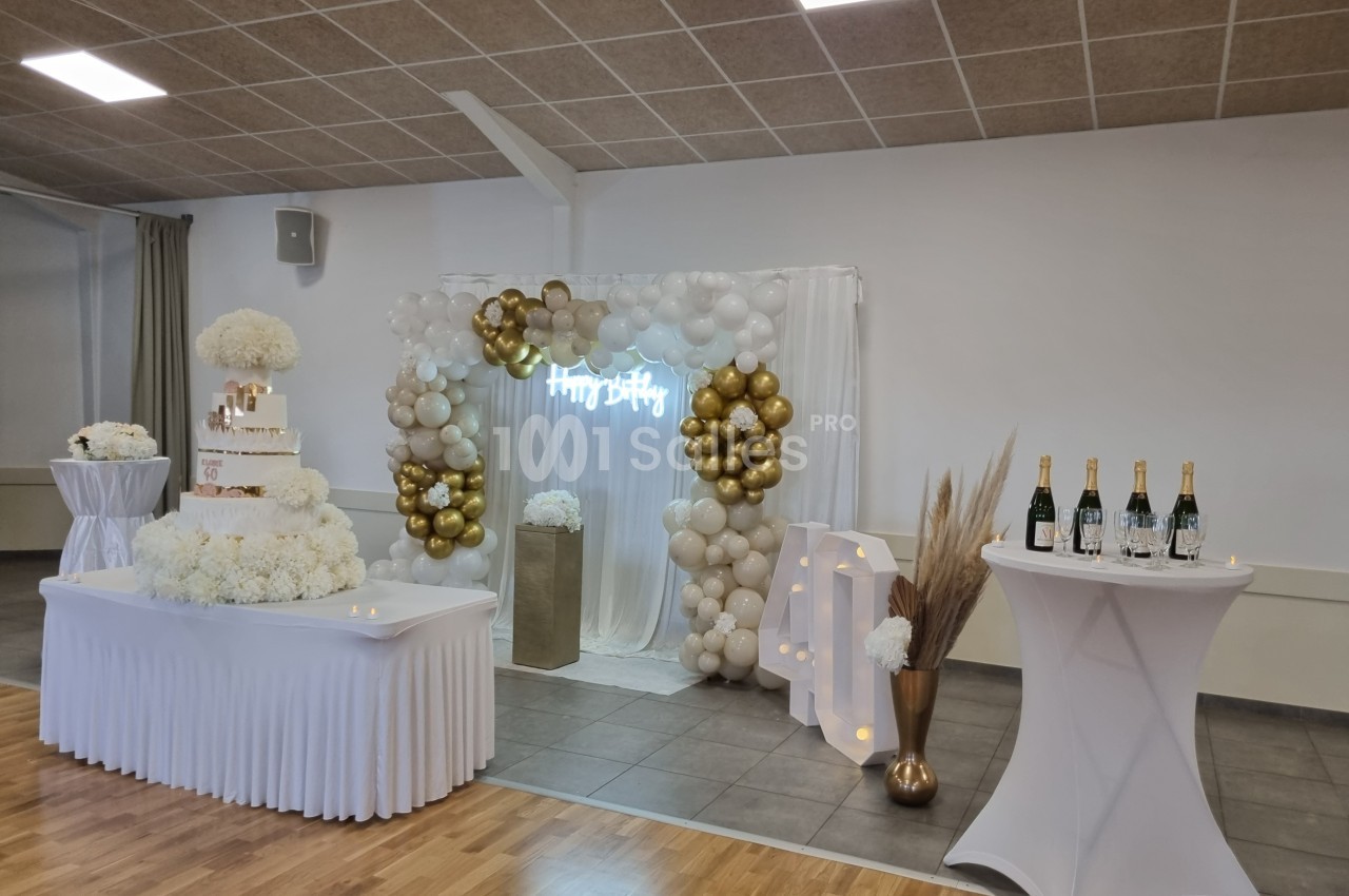 Décoration de fête avec arche de ballons dorés et blancs, table avec gâteau et bouteilles sur des tables hautes.
