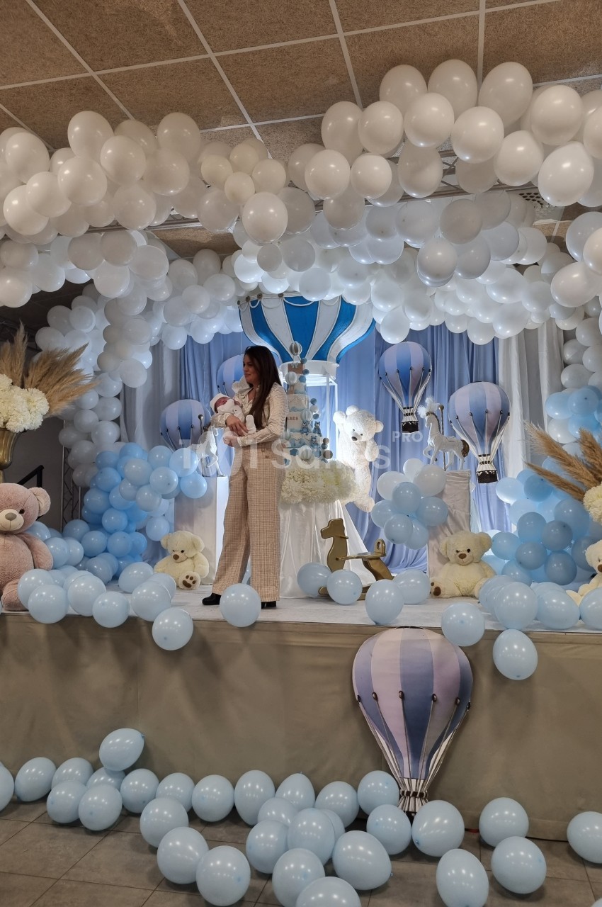 Une femme tient un bébé sur une scène décorée de ballons blancs et bleus, avec des peluches et des montgolfières.