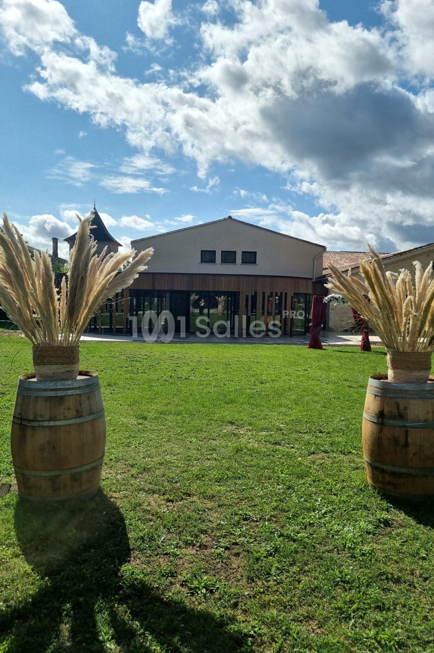 Deux tonneaux décorés de pampas sur une pelouse, devant un bâtiment moderne sous un ciel partiellement nuageux.