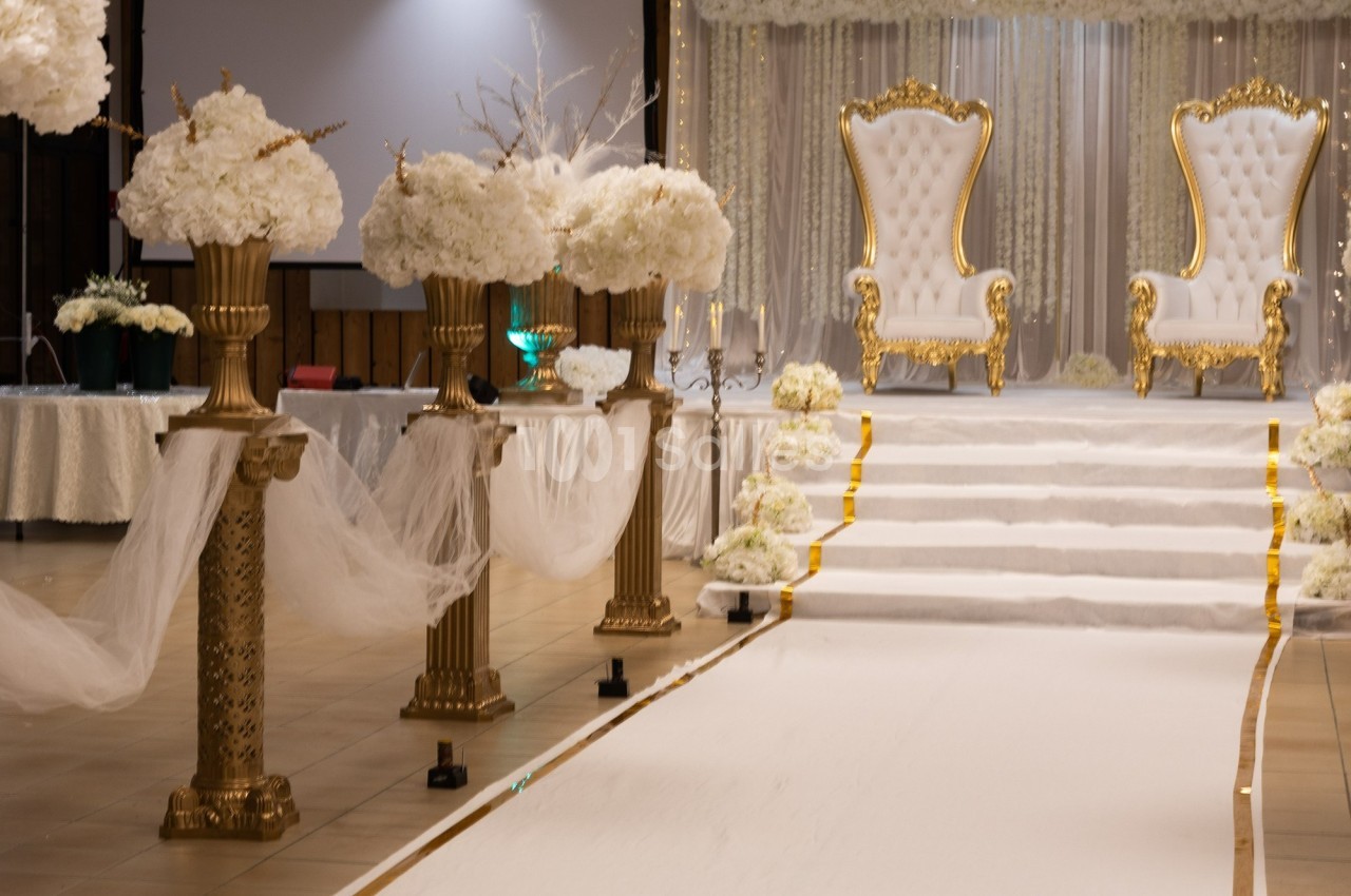 Allée décorée de fleurs blanches menant à une estrade avec deux fauteuils dorés dans une salle de réception.