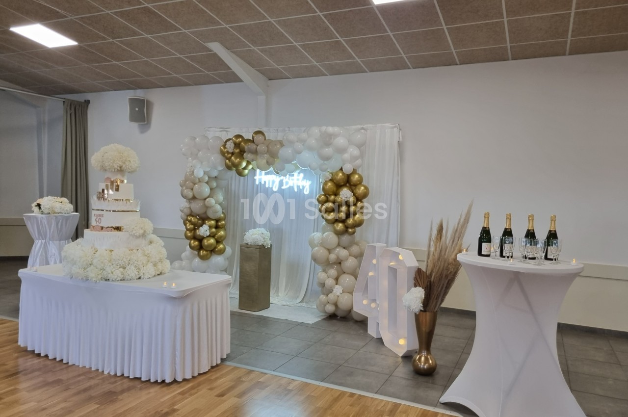 Décoration de salle de fête avec arche de ballons blancs et dorés, gâteau sur une table et bouteilles de champagne.