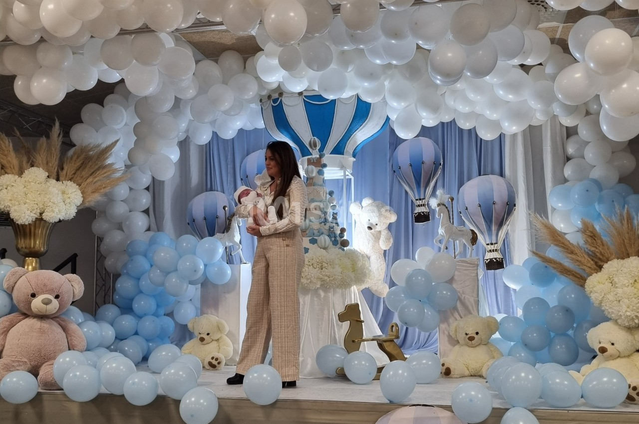 Une femme tenant un bébé dans une salle décorée de ballons bleus et blancs, avec des peluches et des montgolfières.