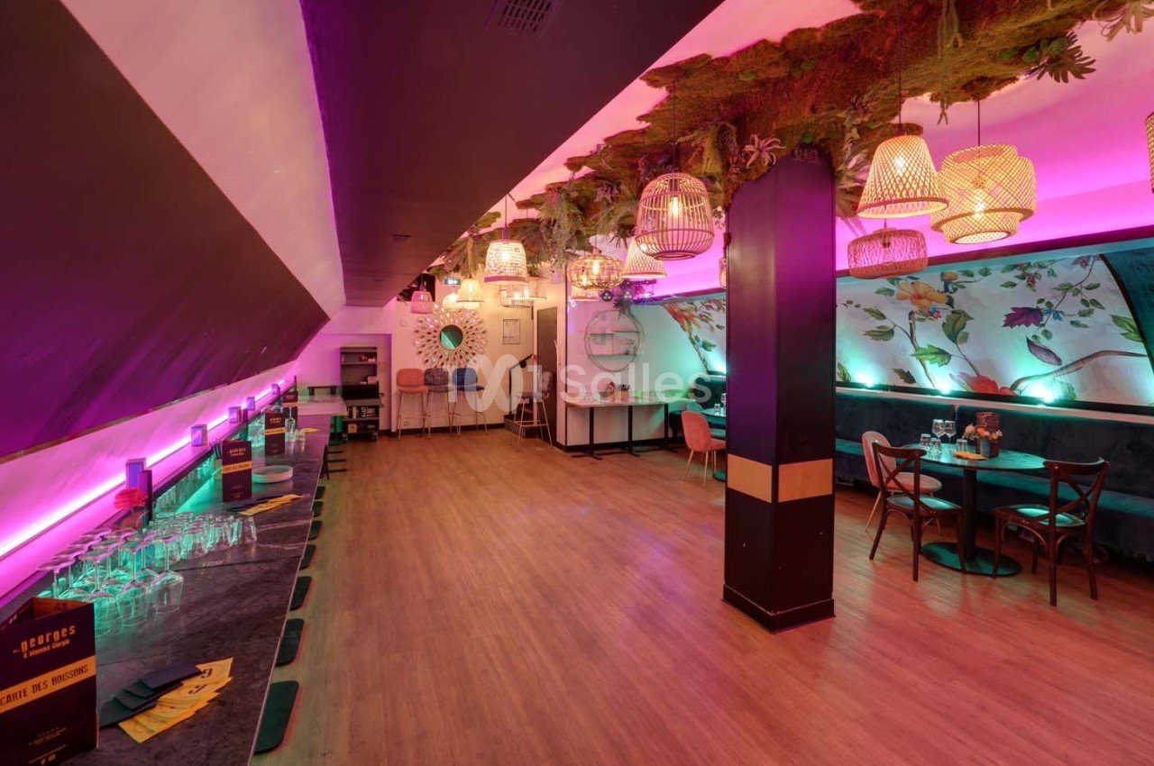 Salle lumineuse avec décoration végétale suspendue, tables et chaises colorées, et un bar sur la gauche.