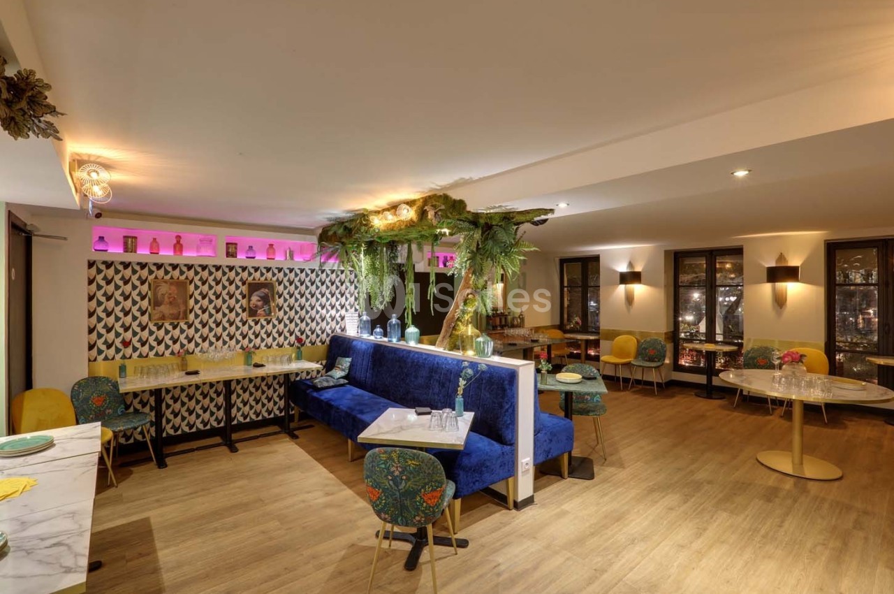 Salle de restaurant lumineuse avec banquettes bleues, chaises colorées, décor végétal et éclairage chaleureux.