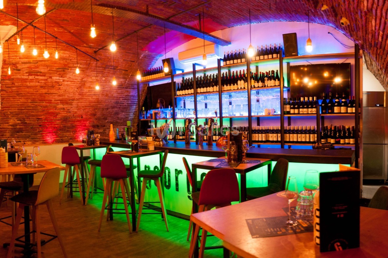 Intérieur d'un bar avec un comptoir lumineux, des étagères de bouteilles et des tables en bois sous un plafond voûté.