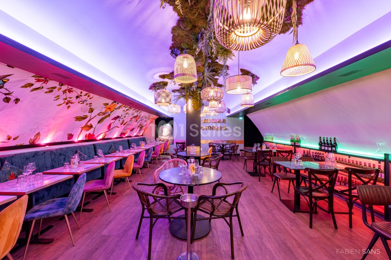 Salle de restaurant colorée avec tables dressées, chaises variées, éclairage tamisé et décoration végétale suspendue.