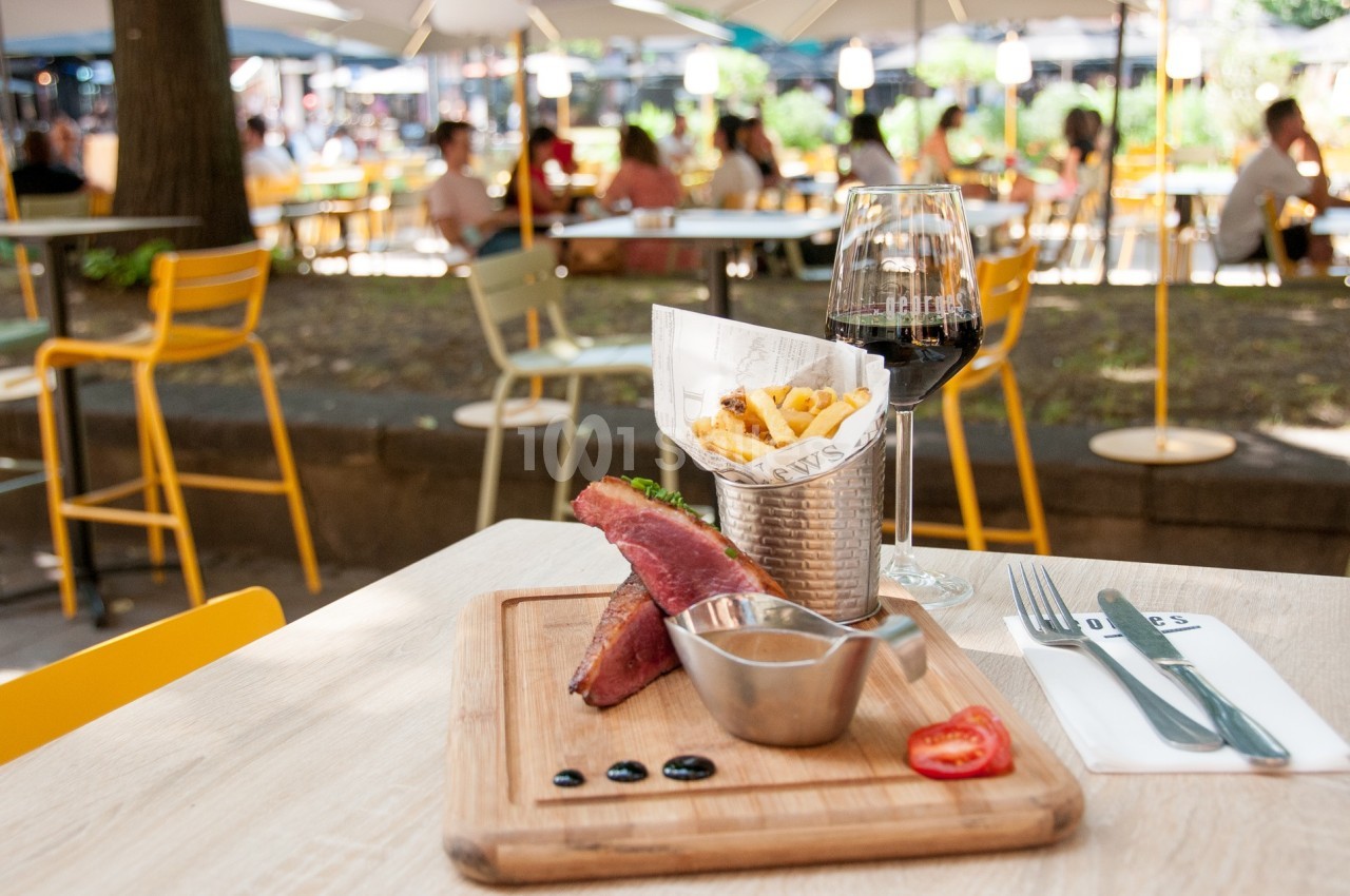 Assiette avec sandwich, frites dans un contenant métallique, sauce, verre de vin rouge, en terrasse ensoleillée.