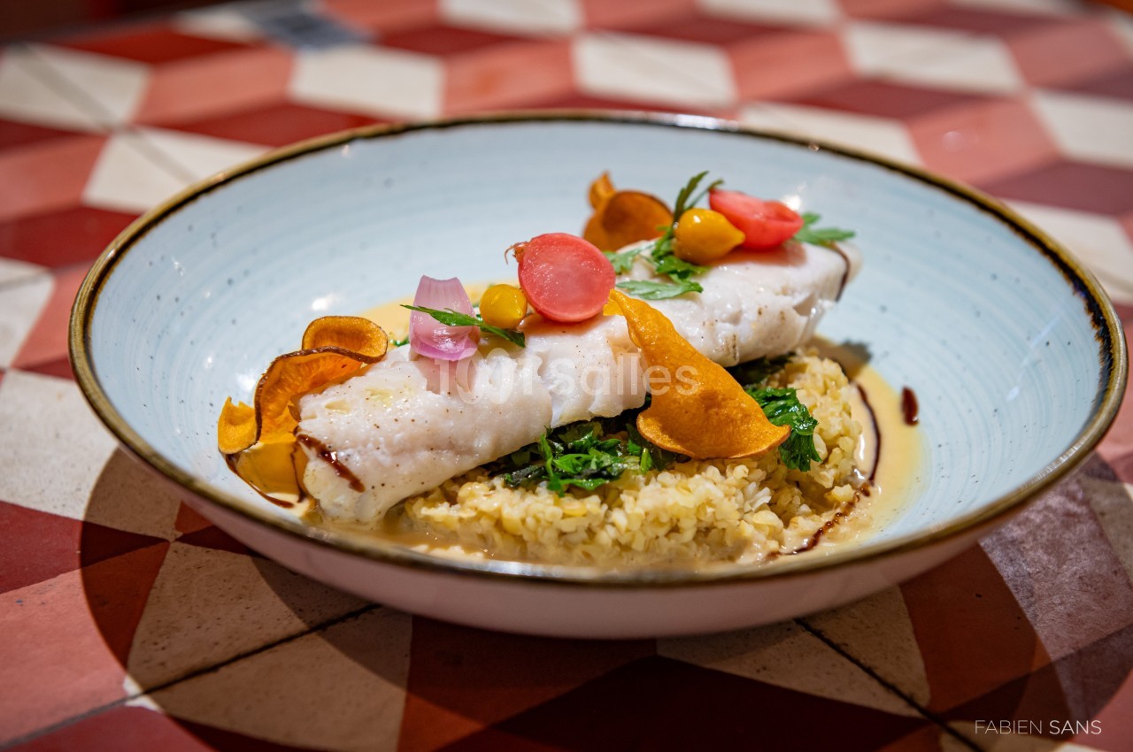 Filet de poisson blanc sur lit de boulgour, garni de légumes colorés et chips de patate douce, dans une assiette creuse.