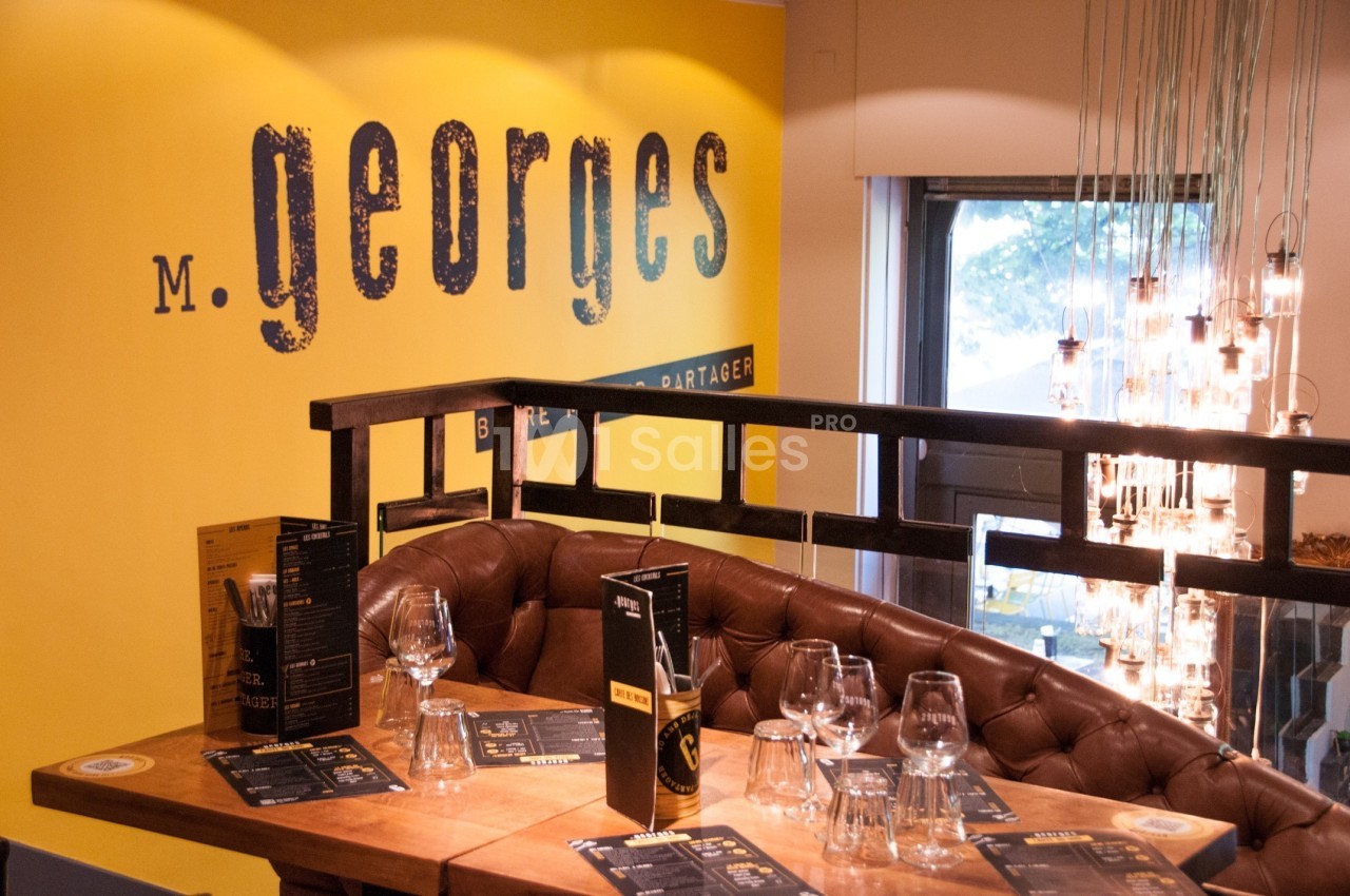Banquette en cuir autour d'une table dressée avec des menus et des verres, devant un mur jaune marqué ’M. Georges’.