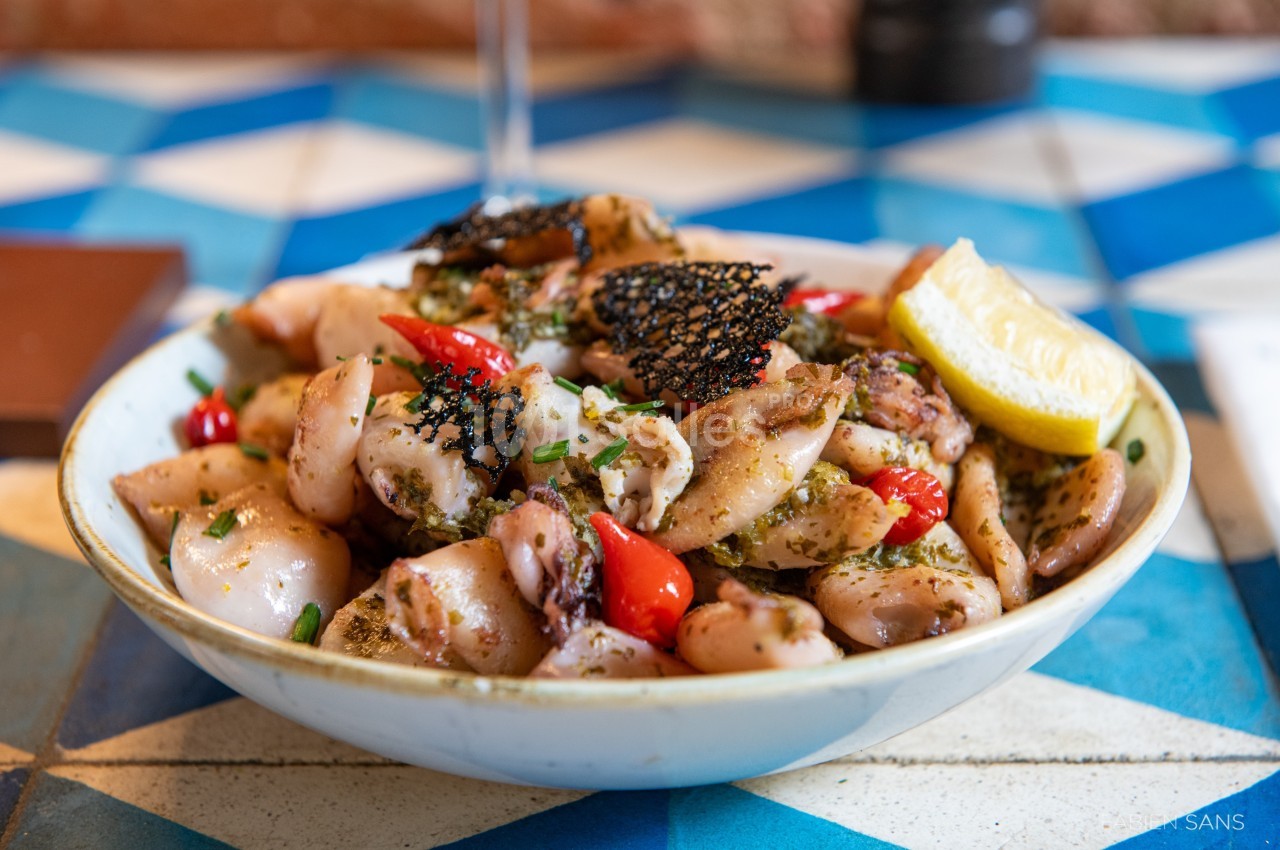 Assiette de calamars grillés garnis de tomates cerises, herbes fraîches et tranche de citron sur une table carrelée.