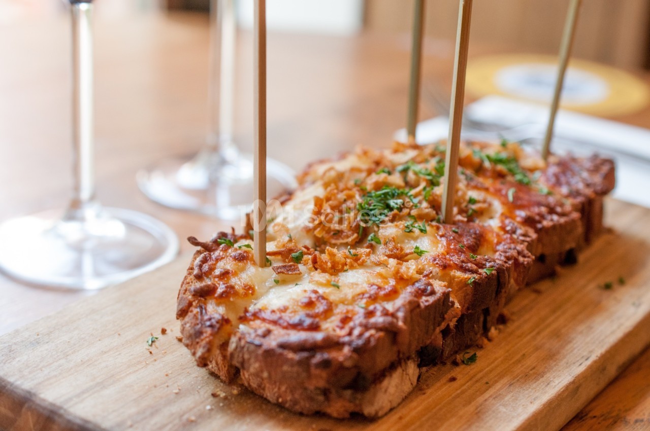 Tranche de pain gratinée au fromage, garnie d'oignons frits et d'herbes, servie sur une planche en bois.