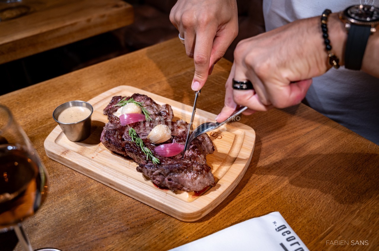 Une personne découpe une pièce de viande garnie d'oignons et d'herbes sur une planche en bois, avec une sauce à côté.