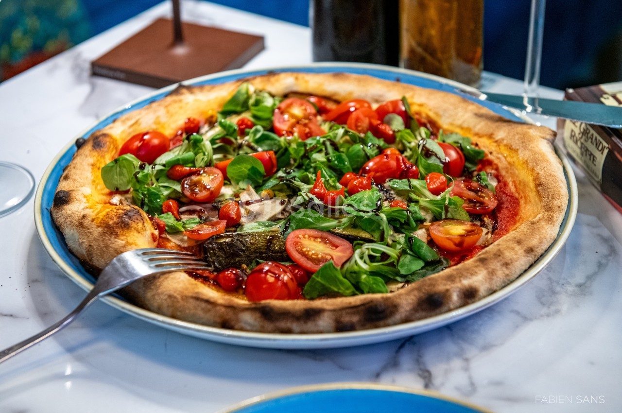 Pizza garnie de roquette, tomates cerises et légumes grillés, servie sur une assiette bleue posée sur une table en marbre.