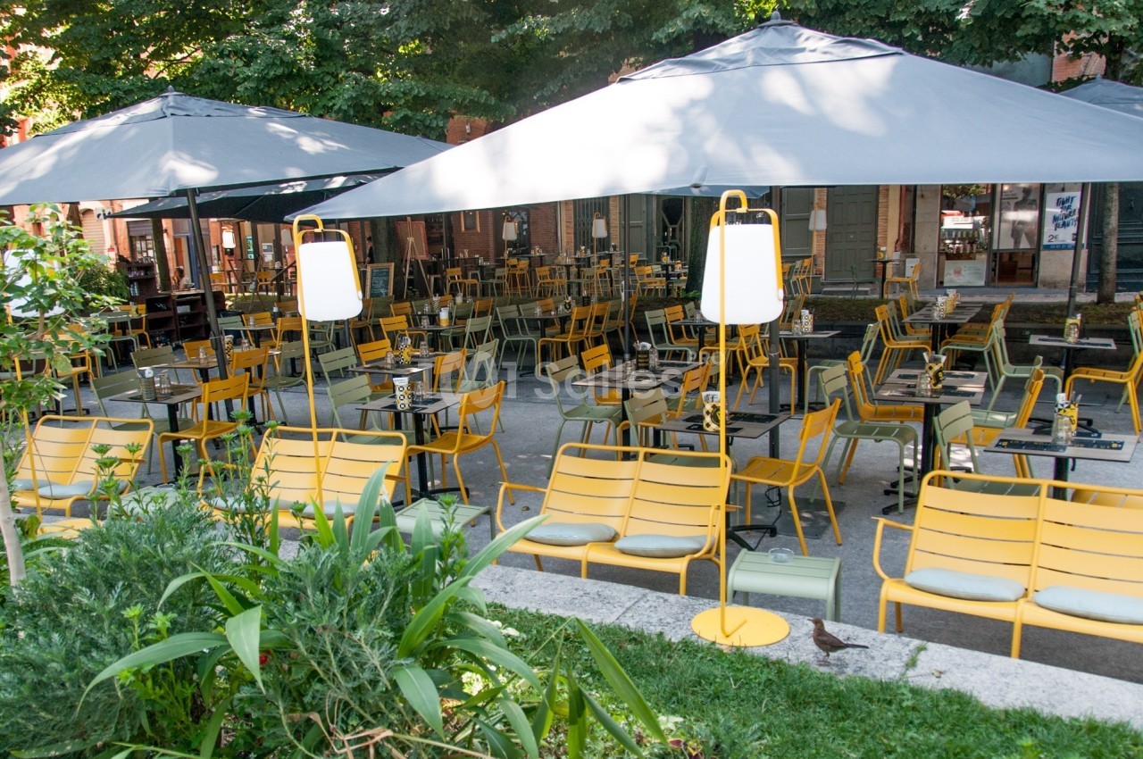Terrasse extérieure avec des tables et des chaises jaunes sous de grands parasols gris, entourée de verdure.