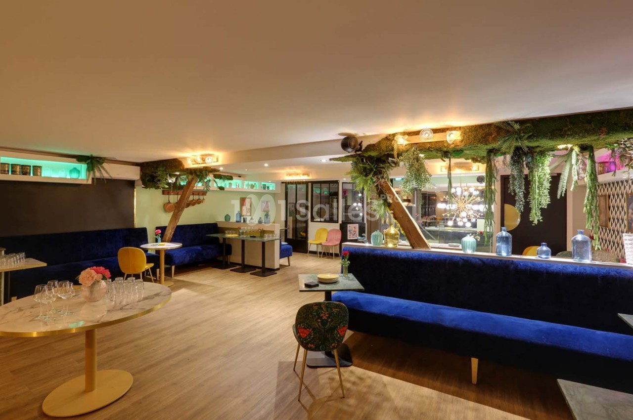 Salle lumineuse avec banquettes bleues, tables en bois clair, décor végétal suspendu et grandes baies vitrées.