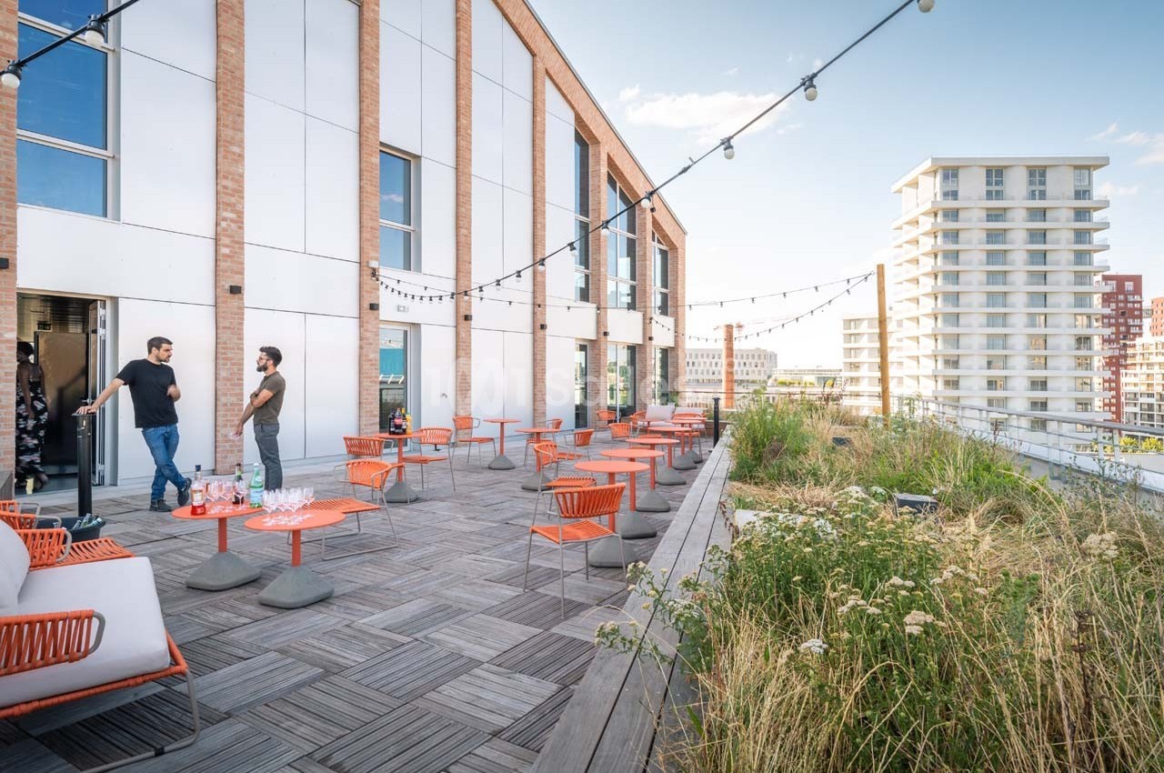 Terrasse en bois aménagée avec tables et chaises, entourée de végétation, donnant sur des immeubles modernes.