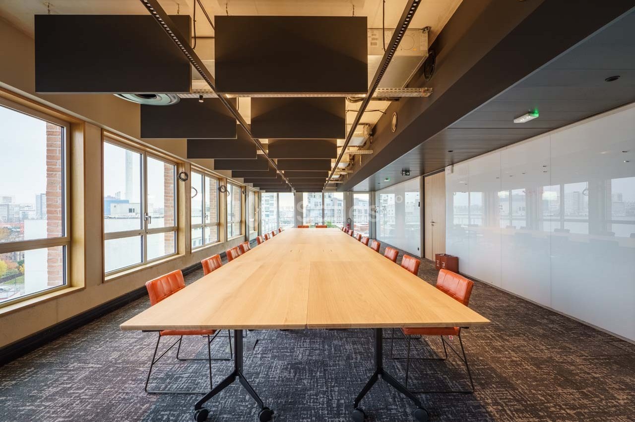 Salle de réunion lumineuse avec une grande table en bois, chaises orange et baies vitrées donnant sur la ville.