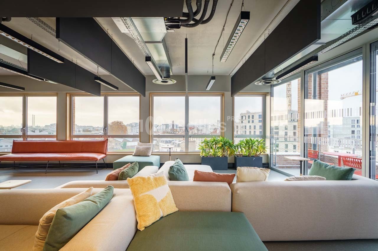 Espace lumineux avec canapés modernes, grandes fenêtres donnant sur une vue urbaine et plantes décoratives.
