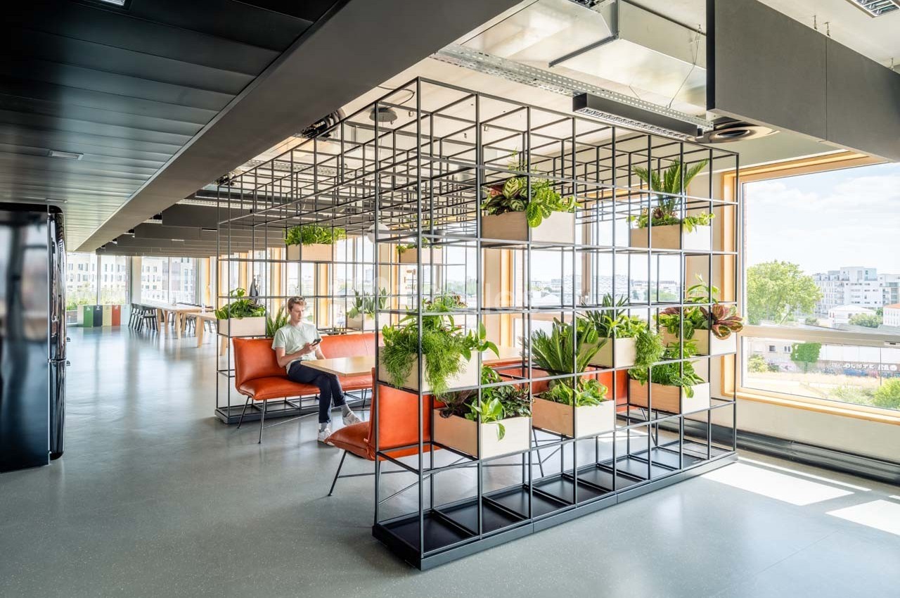 Espace lumineux avec des étagères métalliques remplies de plantes, des sièges orange et une personne assise.