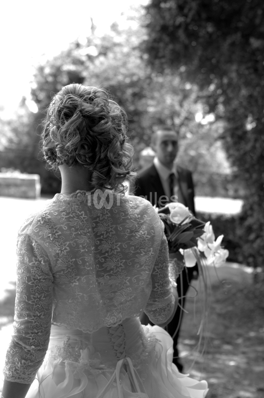 Une mariée de dos tenant un bouquet, face à un homme en costume dans un jardin en noir et blanc.