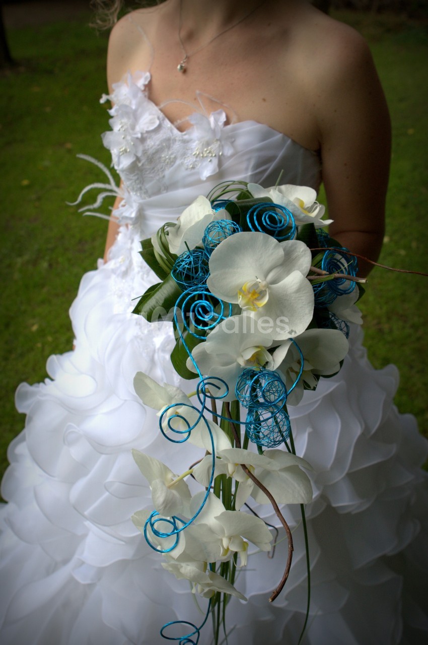 Femme en robe de mariée blanche tenant un bouquet de fleurs blanches et décorations bleues, en extérieur sur pelouse.