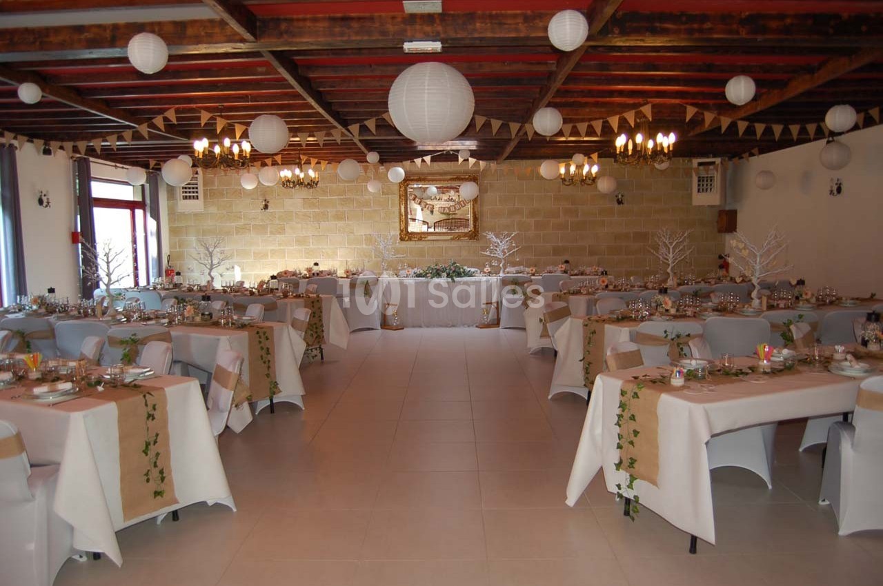 Salle de réception décorée pour un événement, avec tables dressées, guirlandes et lanternes suspendues au plafond.