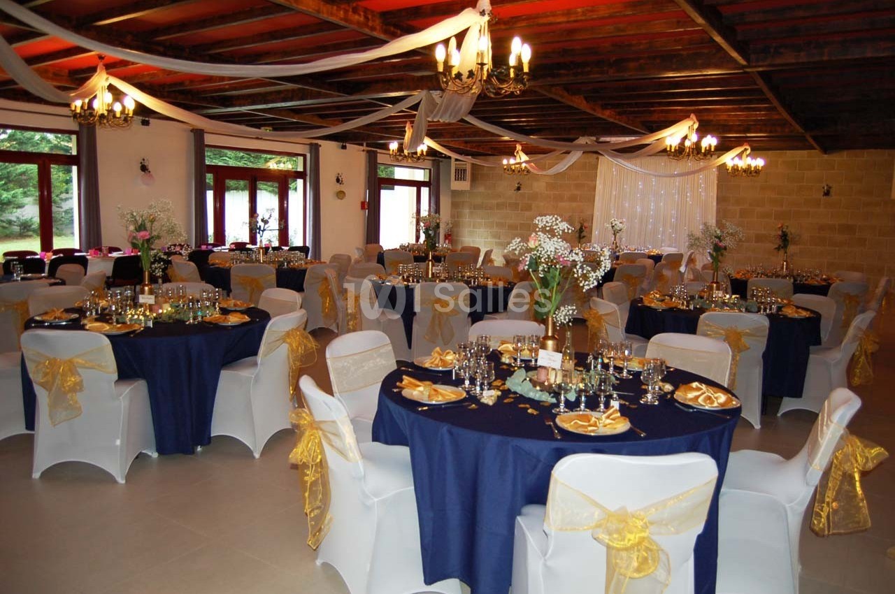 Salle de réception décorée avec des tables rondes nappées de bleu, chaises blanches ornées de nœuds dorés et éclairage…