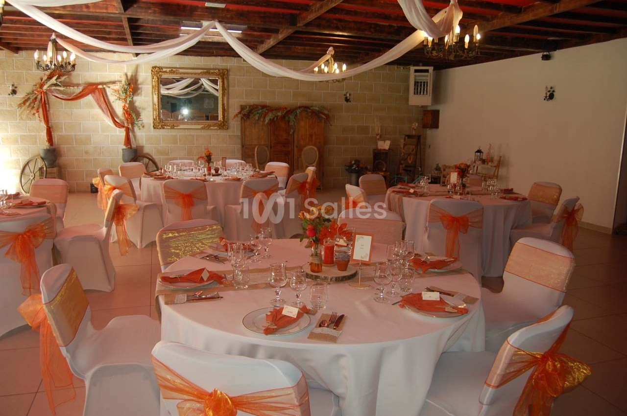 Salle de réception décorée avec des tables rondes, nappes blanches et chaises ornées de rubans orange.