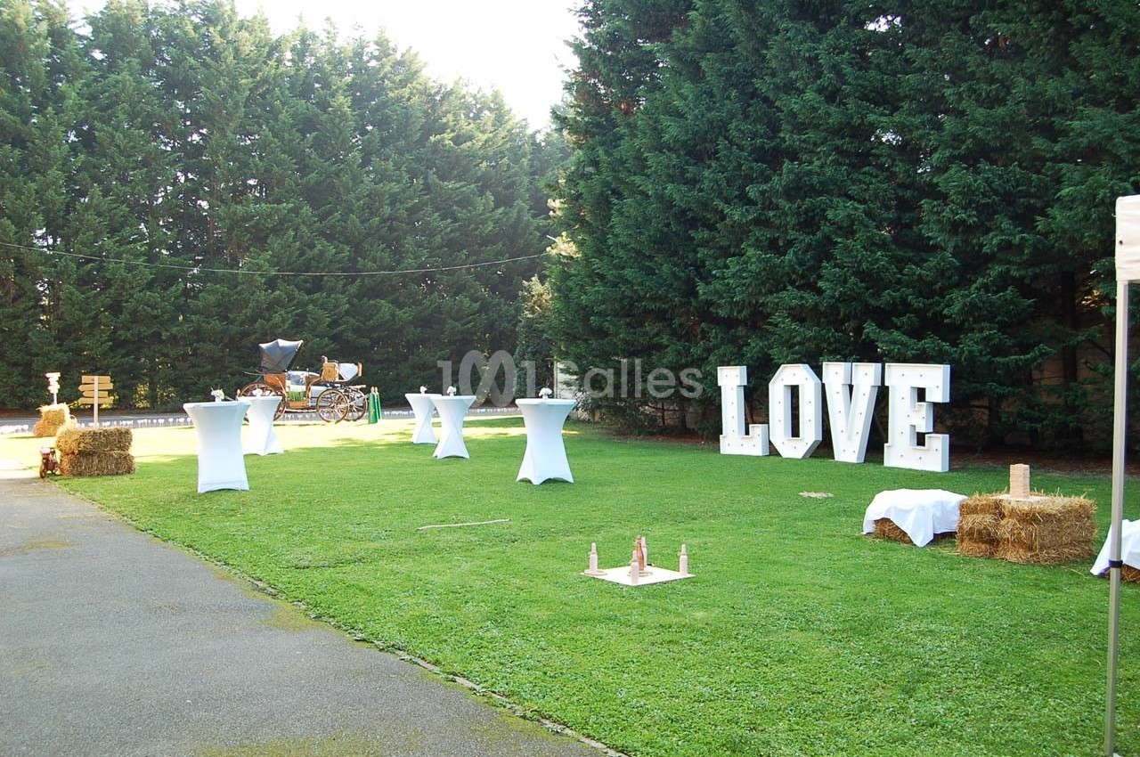 Espace extérieur décoré pour un événement avec lettres lumineuses ’LOVE’, tables hautes et bottes de foin sur une pelouse.