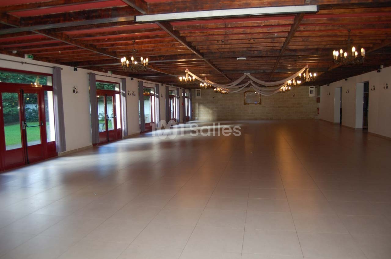 Salle spacieuse avec sol carrelé, plafond en bois, grandes fenêtres et éclairage suspendu, donnant sur un espace vert.