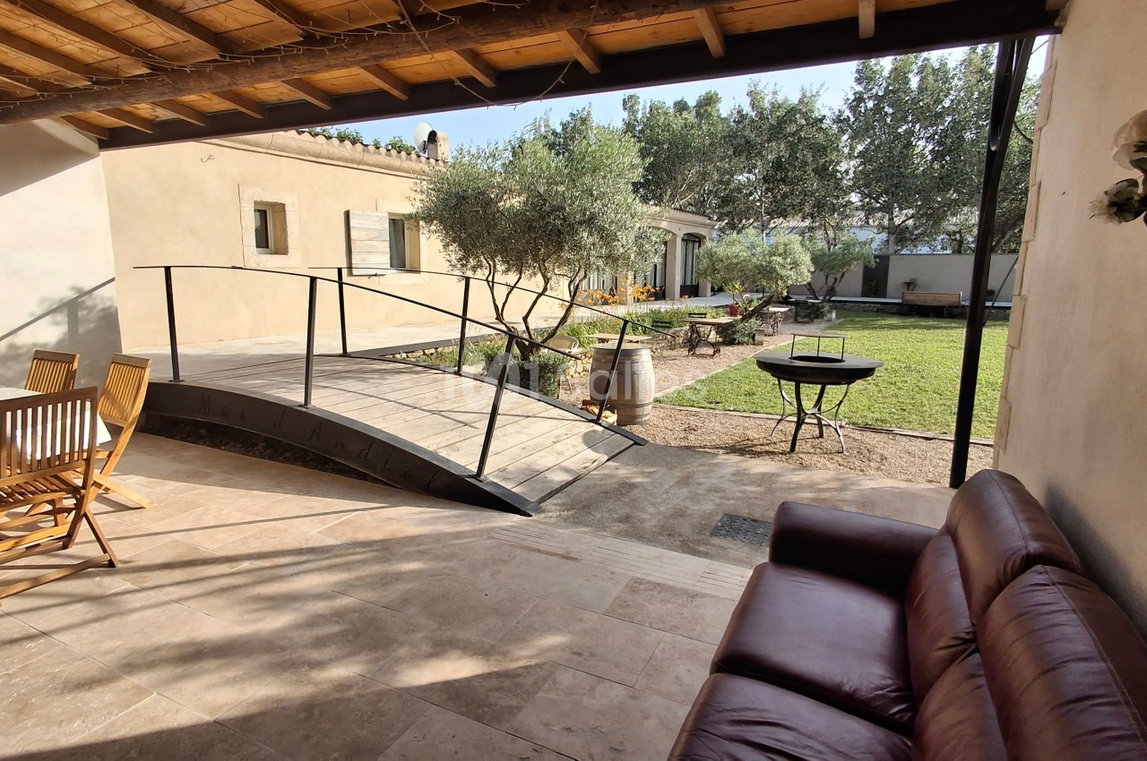 Terrasse avec salon de jardin, canapé, rampe en bois menant à un jardin arboré avec barbecue et oliviers.