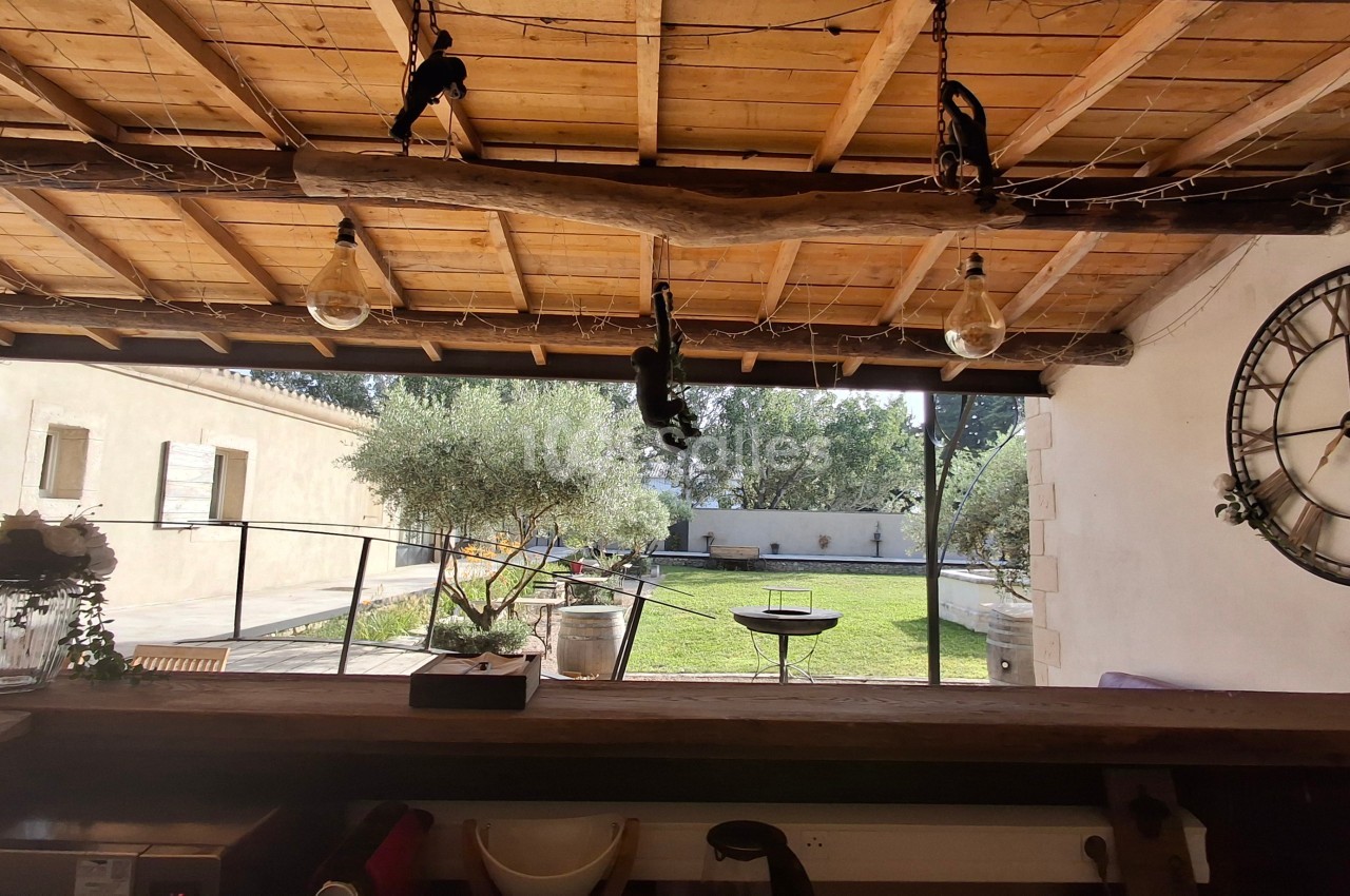 Vue depuis un bar en bois sous une pergola, donnant sur un jardin avec olivier, barbecue et horloge murale.